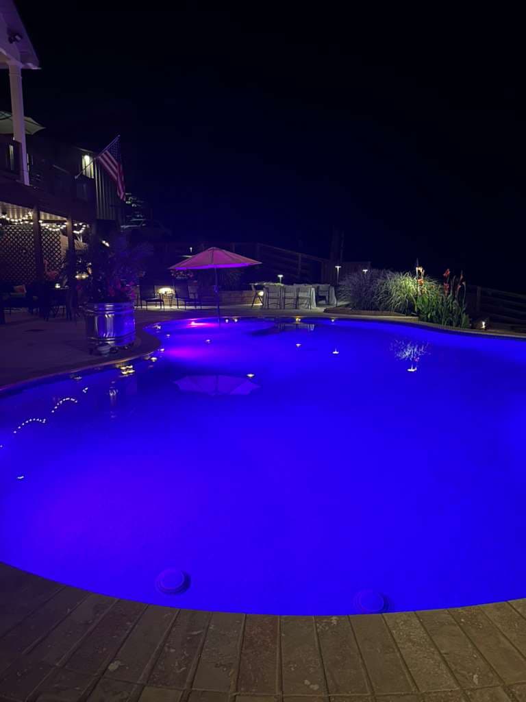 Illuminated backyard pool with purple lights and seating area at night.