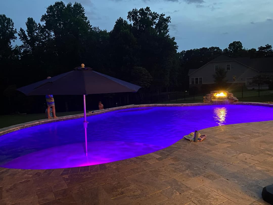 Evening view of a pool with purple lighting, umbrella, and children playing nearby.