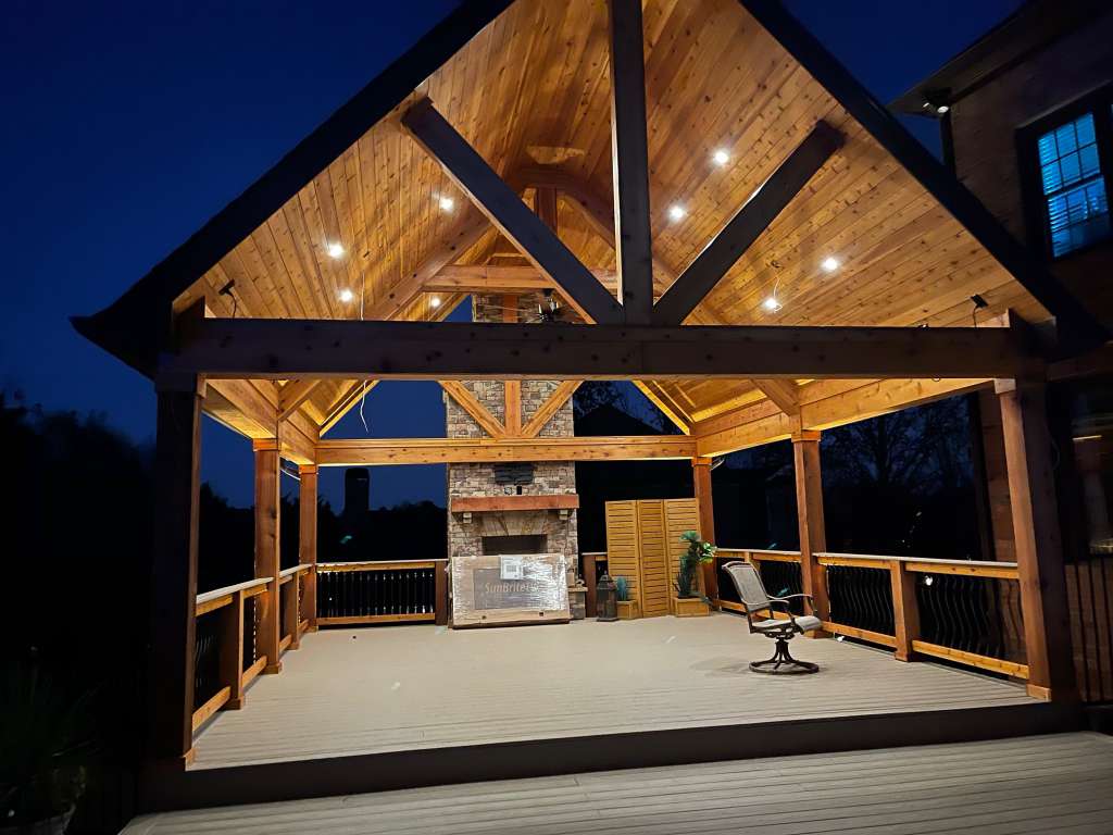 Elegant wooden gazebo at night with ambient lighting and stone fireplace, perfect for gatherings.