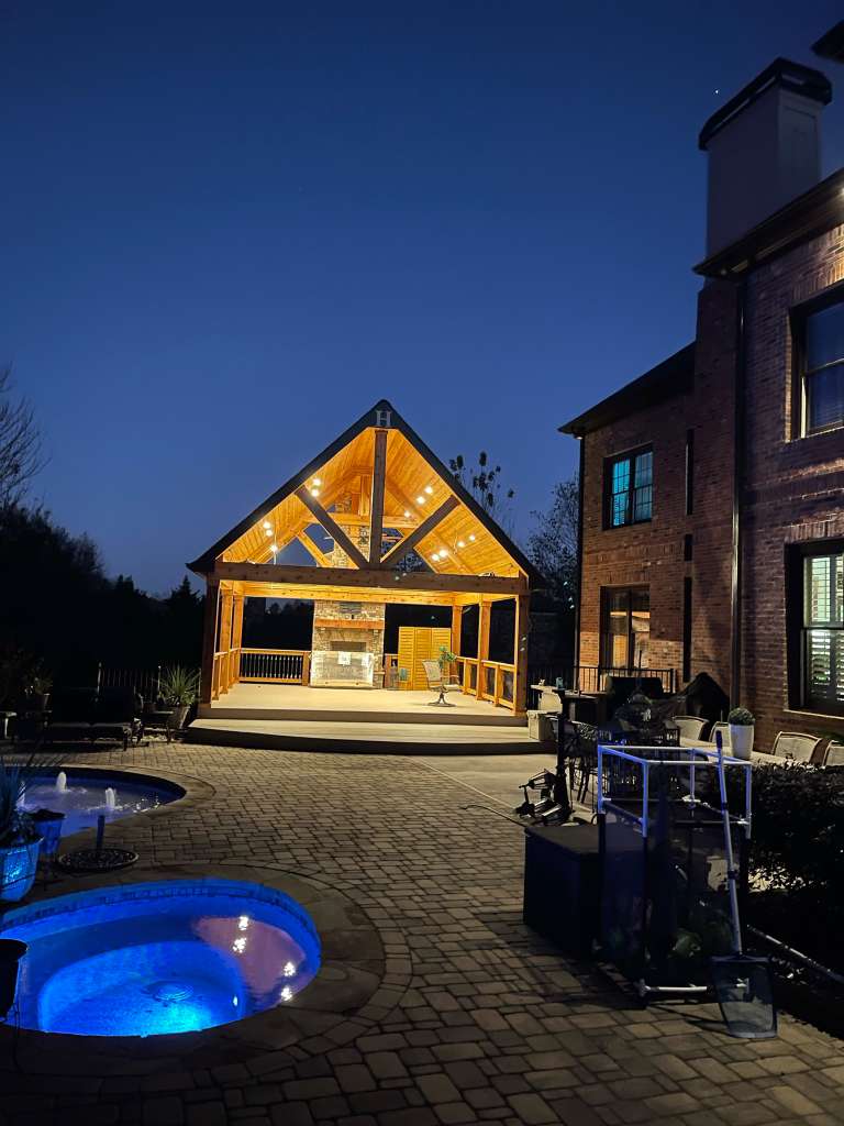 Modern outdoor pavilion with wooden beams, lit at night, overlooking a patio and pool.