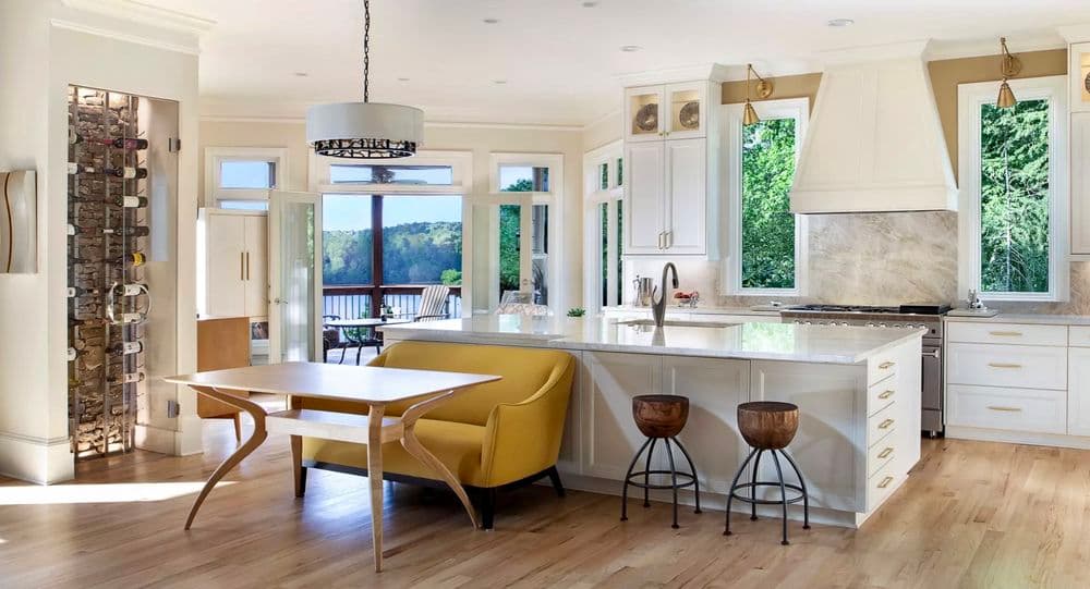 Modern kitchen with white cabinetry, yellow dining table, bar stools, and large windows overlooking nature.