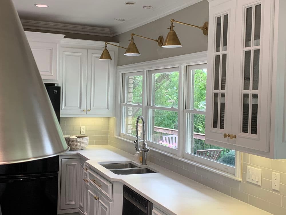 Modern white kitchen with brass fixtures, large windows, and sleek countertops. Natural light.