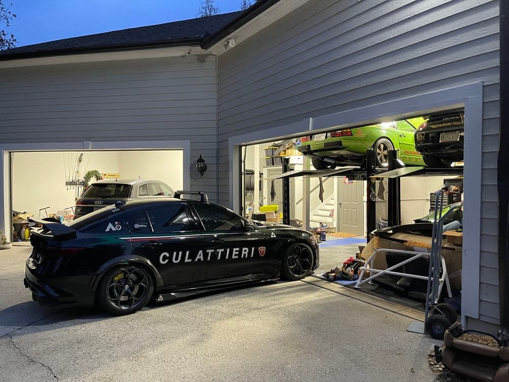 Black sedan in driveway next to garage with lifted green car and tools, evening setting.