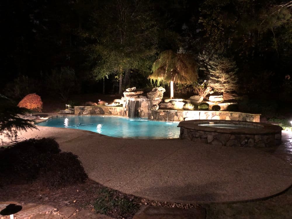 Illuminated backyard pool with waterfall and stone features at night, surrounded by lush greenery.