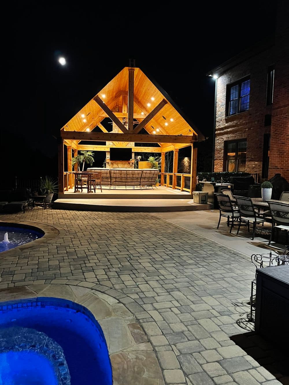Cozy outdoor gazebo with lighting by a pool at night, surrounded by stone patio and seating.